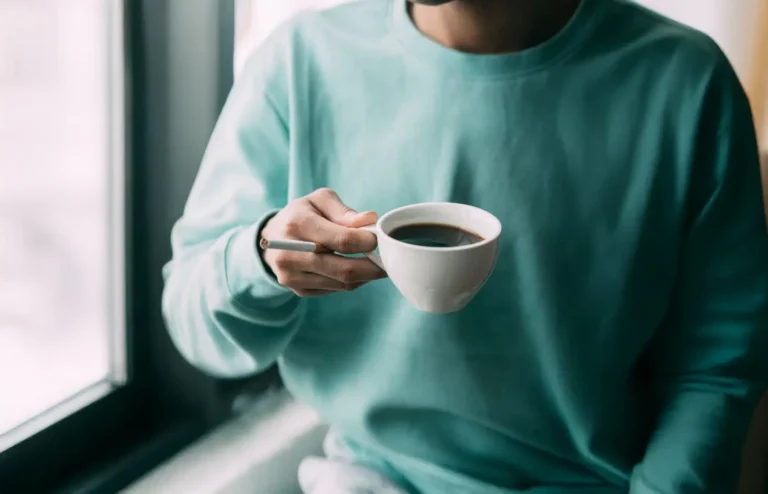 Personne tenant une tasse de café, profitant d’un moment de détente à l’intérieur, vêtue d’un pull vert d’eau.