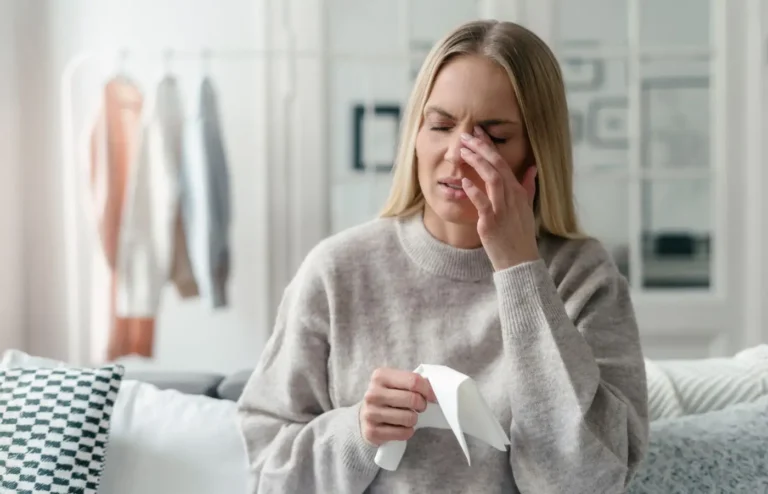 Femme se frottant les yeux avec une expression de gêne, tenant un mouchoir, illustrant les symptômes de sécheresse oculaire ou d'irritation causés par des facteurs externes comme la fumée.