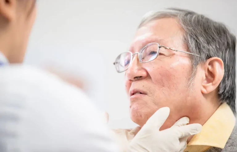 Un médecin examine la gorge d'un client âgé, illustrant une consultation pour des problèmes vocaux ou respiratoires potentiellement causés par le tabagisme.