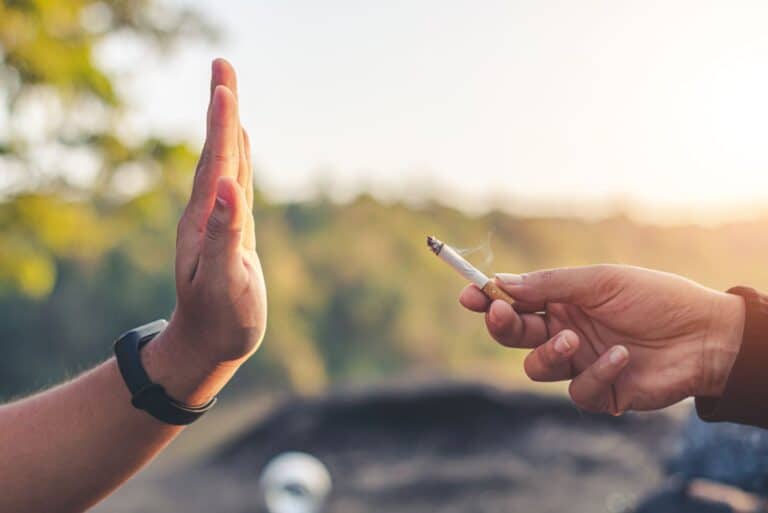 Gros plan sur deux mains sur fond de paysage extérieur. La main de droite propose une cigarette allumée à la main de gauche, qui la refuse d'un geste catégorique.