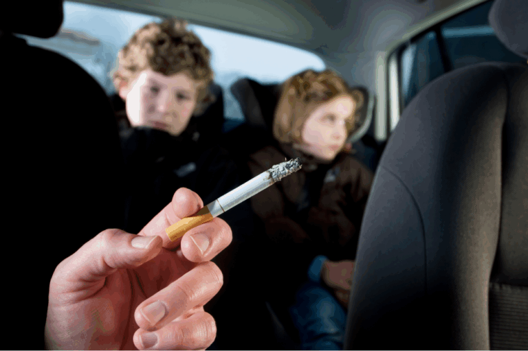 Gros plan d'une main tenant une cigarette allumée dans l'habitcle d'une voiture, deux jeune enfants sur les sièges arrières.