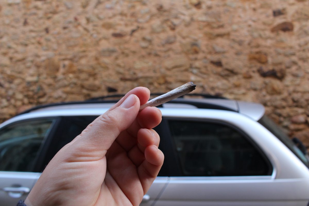 Main tenant un joint de cannabis éteint, devant une voiture à l'arrêt