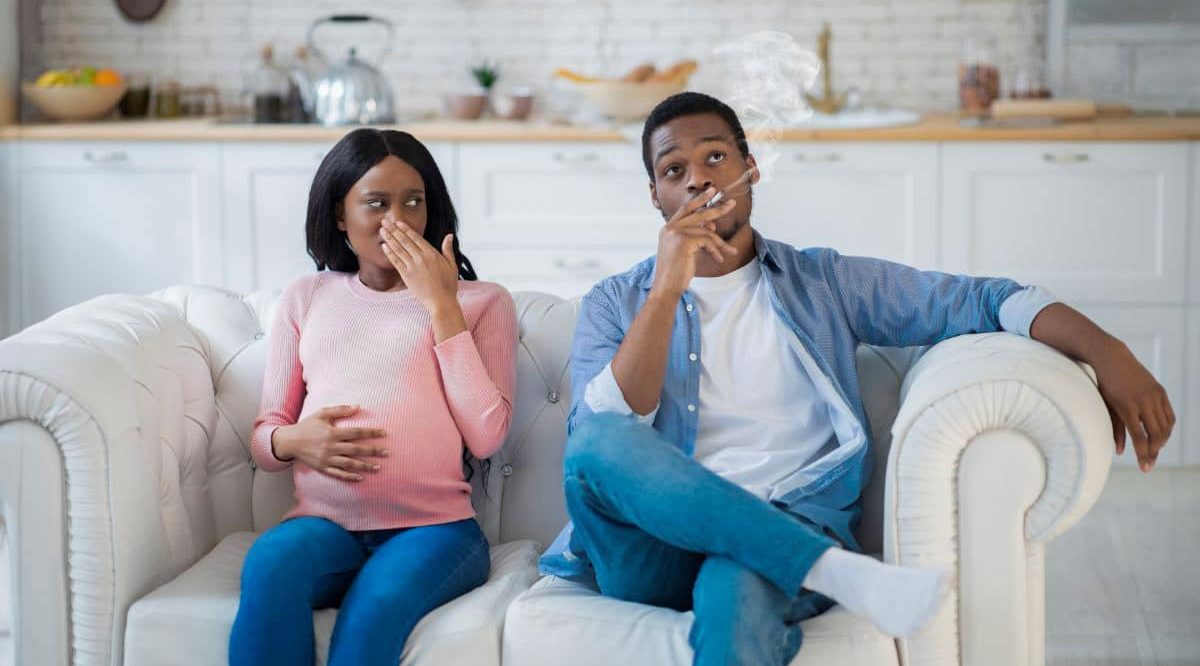 Couple assis sur un canapé, la femme est enceinte et se touche le nez car l'homme fume juste à côté d'elle.