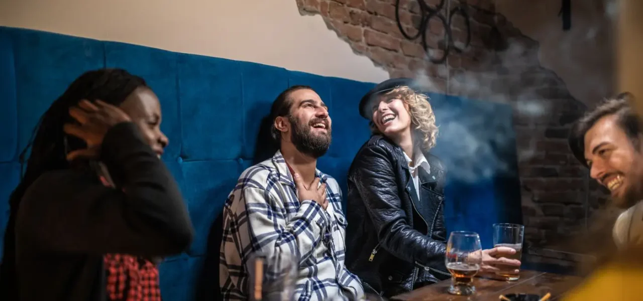 Un groupe d’amis assis dans un bar, partageant des verres et des rires, avec une personne fumant en arrière-plan.