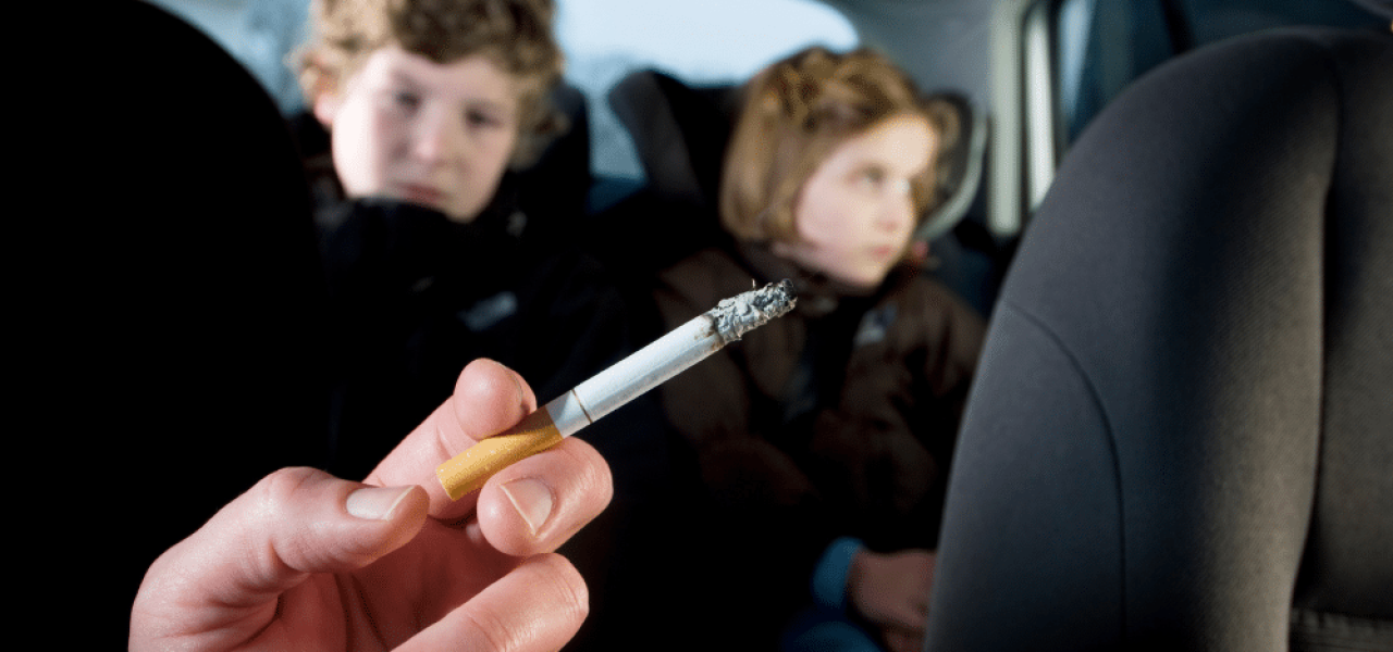 Gros plan d'une main tenant une cigarette allumée dans l'habitcle d'une voiture, deux jeune enfants sur les sièges arrières.